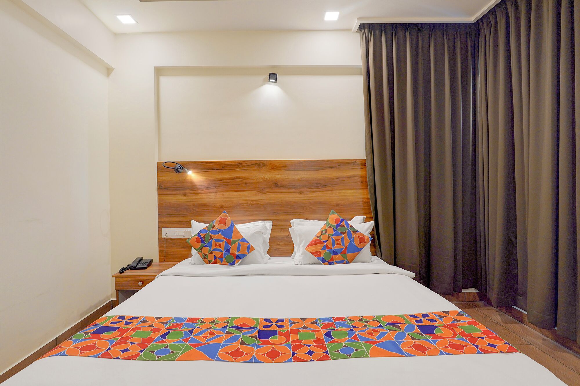 A hotel room with a wooden headboard, a bed with white linens, two colorful geometric pillows, a bedside table with a phone, and a large window with dark curtains.