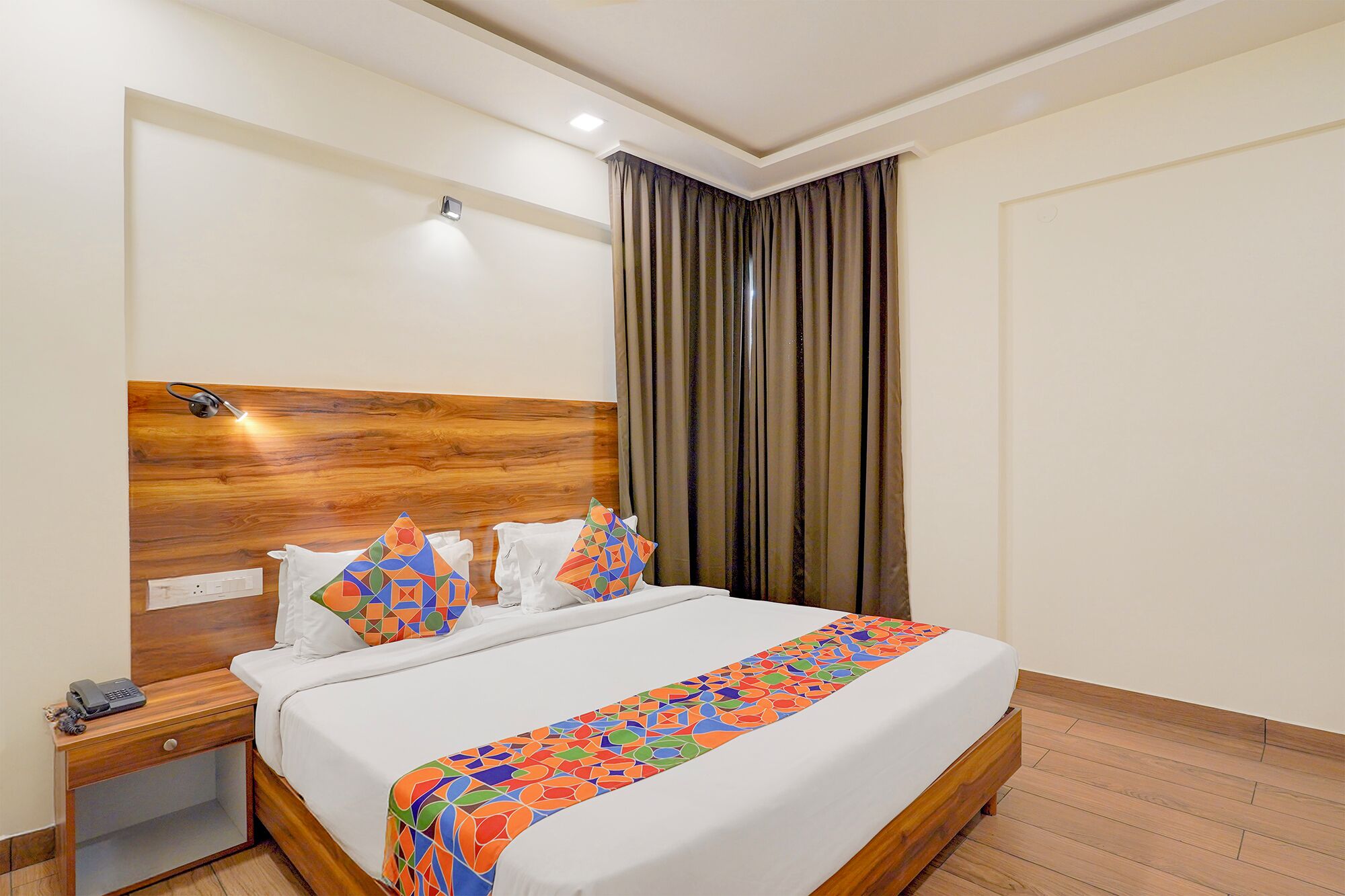 Ein Hotelzimmer mit einem hölzernen Kopfteil, einem Bett mit weißen Leinen, farbenfrohen Kissen, einem Nachttisch mit Telefon und einem Beistelltisch.