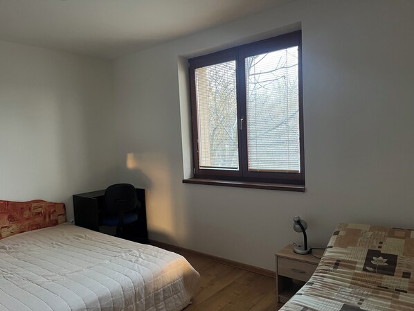 A bedroom with a bed, a desk, a chair, a nightstand, and a window with blinds.