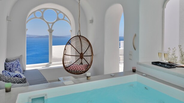 A modern, minimalist room with a large window overlooking the sea, a hanging chair, a small table with a glass, and a bathtub. - Namaste Suites by Oias Local Cavehouses (Oia)