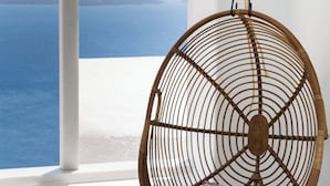 A wicker hanging chair with a cushion, a small table with a pitcher and glasses, and a view of the sea through a white arched window. - Namaste Suites by Oias Local Cavehouses (Oia)