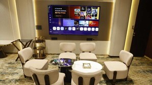 A hotel room with a large flat-screen TV displaying various channels, a seating area with four cushioned chairs, a small round table, and a decorative gold-colored side table. - Siya Residency (New Delhi)