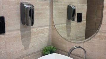 Un baño moderno con un lavamanos blanco, un dispensador de jabón líquido, un dispensador de gel antibacterial, un espejo redondo y una pequeña planta en maceta.