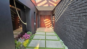 Un estrecho sendero conduce a una puerta de madera, bordeado por un muro de ladrillo negro y un edificio de color oscuro con techo de cristal.