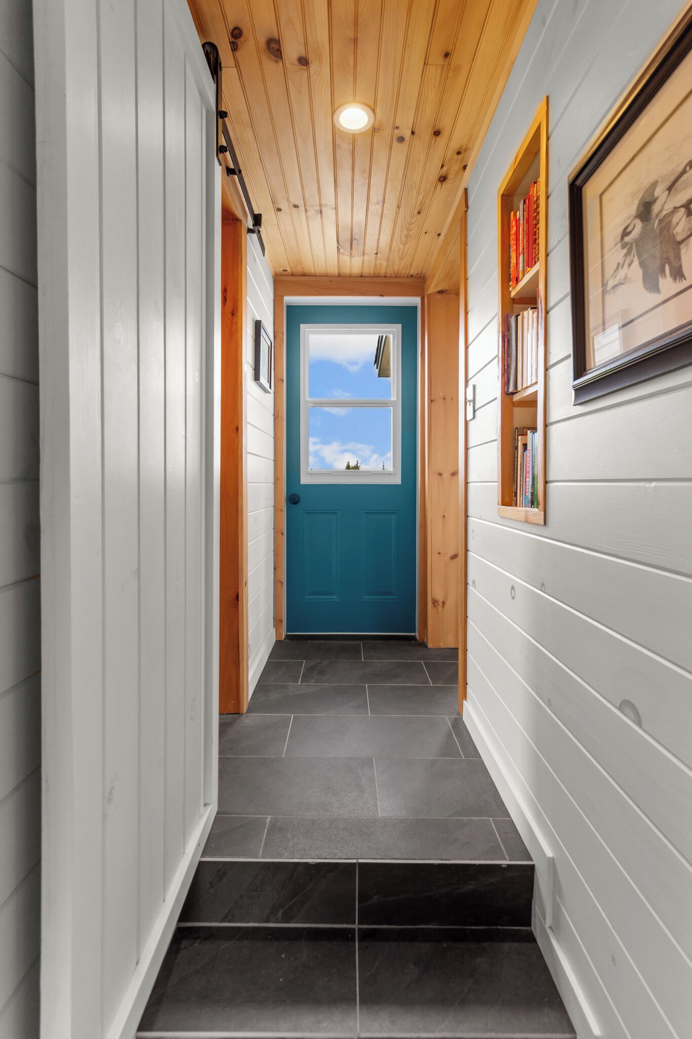 A narrow hallway with wooden ceiling and white walls, leading to a blue door with a window above it.