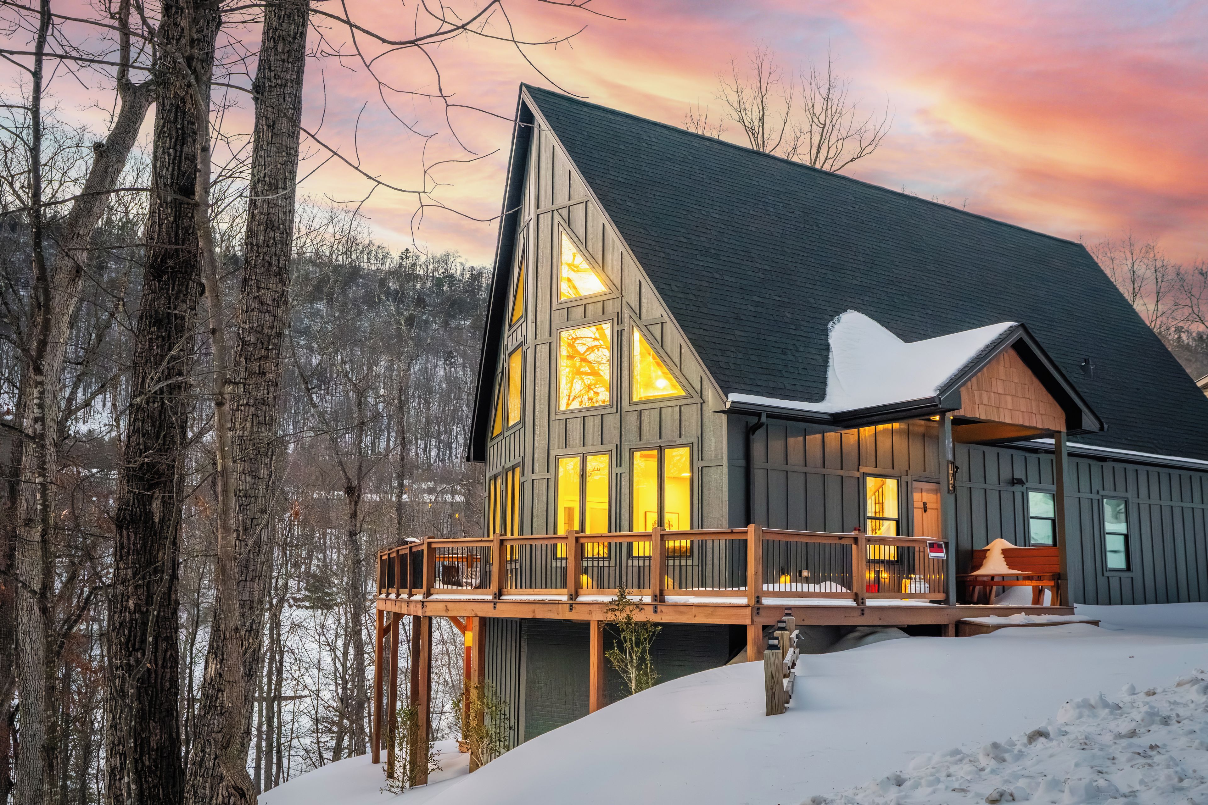 A cabin with a large deck, multiple windows, and a steep roof, surrounded by trees and snow.