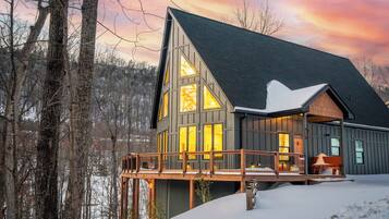 A cabin with a large deck, multiple windows, and a steep roof, surrounded by trees and snow.