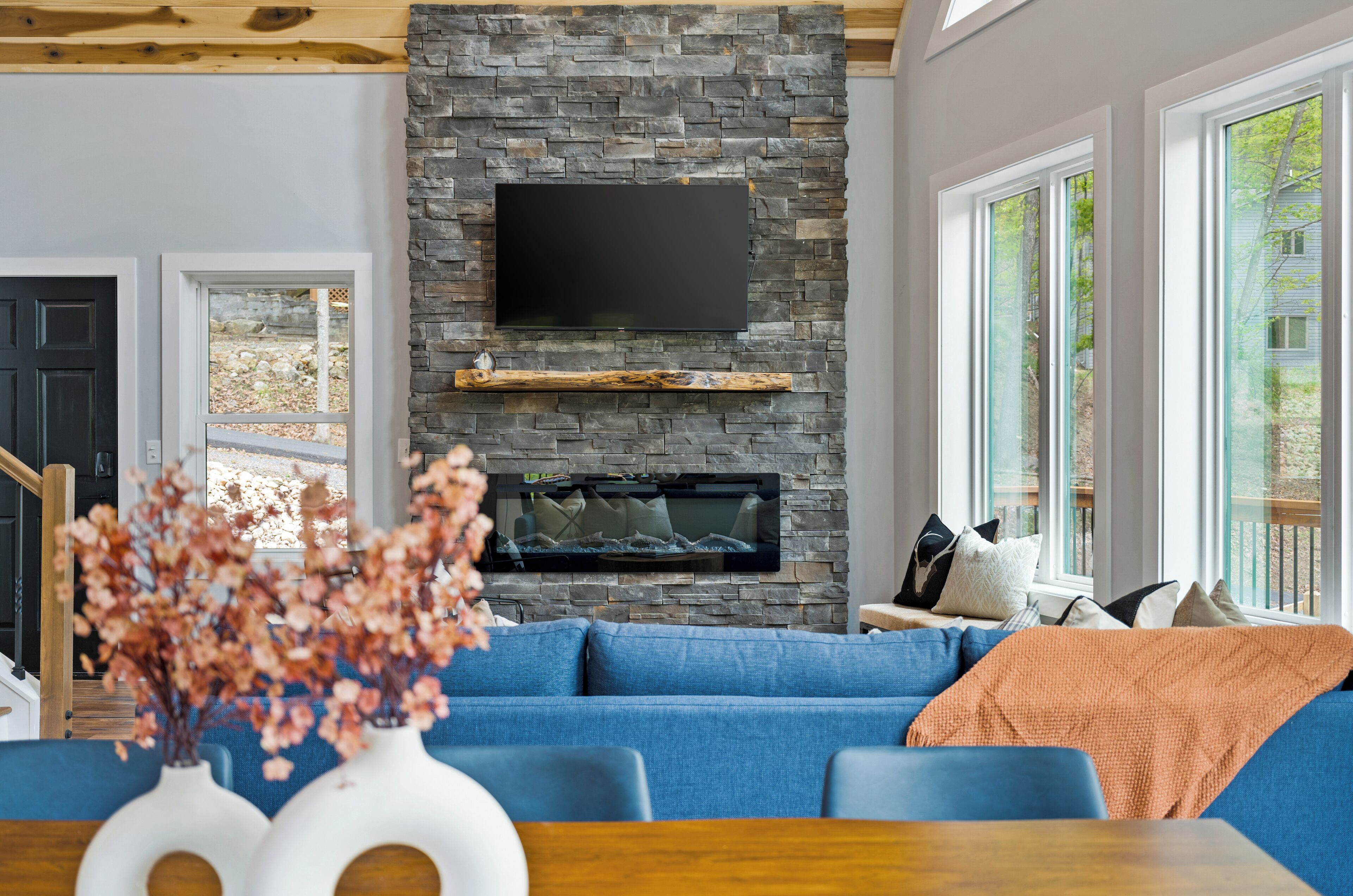 A modern living room with a stone fireplace, a flat-screen TV, and a large window offering a view of the outdoors.