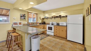 Una cocina con una isla central, taburetes de madera, un cubo de basura, un microondas, un refrigerador y un ventanal con persianas.
