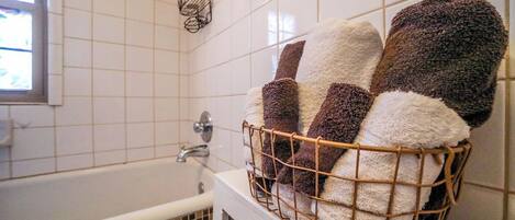 A bathroom with a white bathtub, a towel rack with a wire basket holding several towels, and a window with a view of the outside.