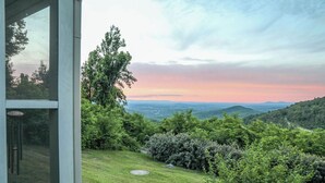 A view from a window overlooking a lush green landscape with a variety of trees and shrubs, and distant hills under a clear sky. - Unwind in a charming historic cottage near Nelson 151 featuring a Level 2 EV outlet, modern comforts, and easy access to Virginia's wineries, breweries, hiking trails, and Blue Ridge scenery. (Afton)