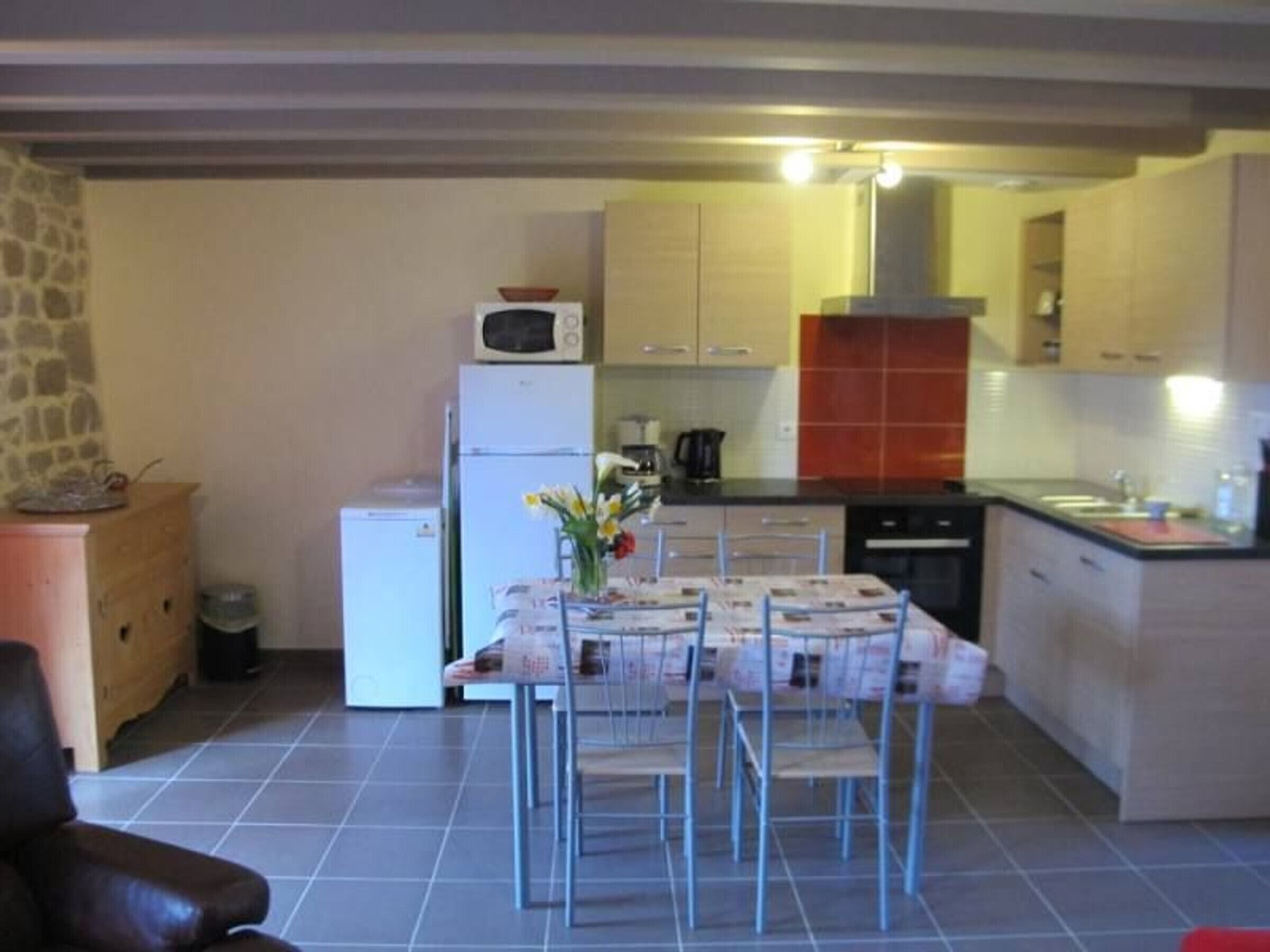 A compact kitchen with a refrigerator, microwave, and stove. There is a dining table with chairs and a vase of flowers. The kitchen has light-colored cabinets and a tiled floor.