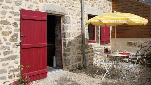 A stone building with red shutters, a small outdoor seating area with a yellow striped umbrella, and a table with chairs.