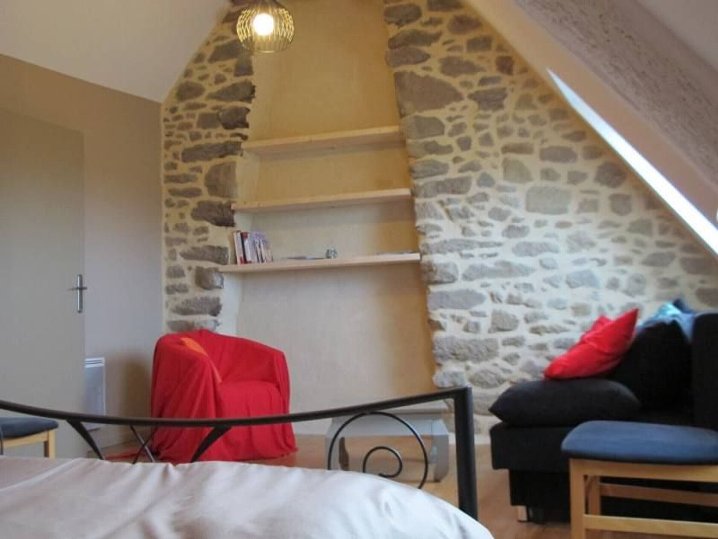Une chambre avec un mur en pierre, un lit, une chaise rouge, un canapé noir et une petite étagère avec des livres.