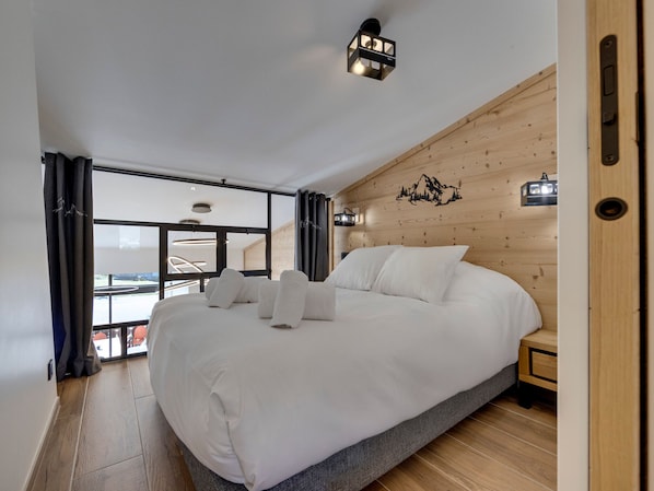 Un dormitorio moderno con una cama grande, paneles de madera en las paredes, vista a través de un ventanal y una lámpara en el techo.