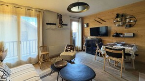 Un salon confortable avec un canapé, deux fauteuils, une table basse et une table à manger avec des chaises. Un téléviseur est fixé au mur, il y a une cheminée et une fenêtre avec des rideaux.