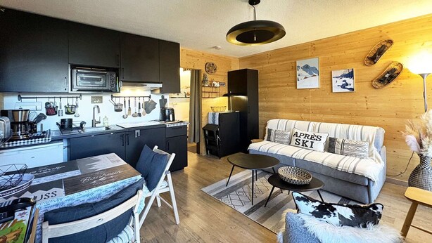 Une cuisine moderne avec des meubles sombres, un four à micro-ondes et une machine à café. On y trouve un coin repas avec une table et des chaises, ainsi qu’un canapé agrémenté d’un coussin sur lequel est inscrit « APRÈS-SKI ».