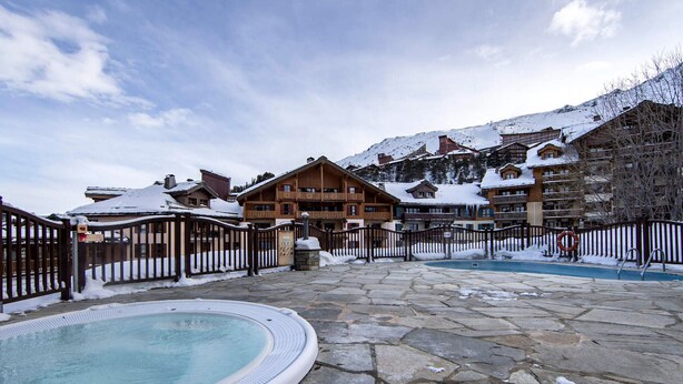A pool area with a hot tub, stone flooring, wooden buildings, and a snowy mountain backdrop.