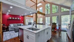 A spacious kitchen with a large island, a dining area with a red wall, and large windows offering a view of the outdoors. - Legends Lodge: A woodland retreat near University of Alabama! (Cottondale)