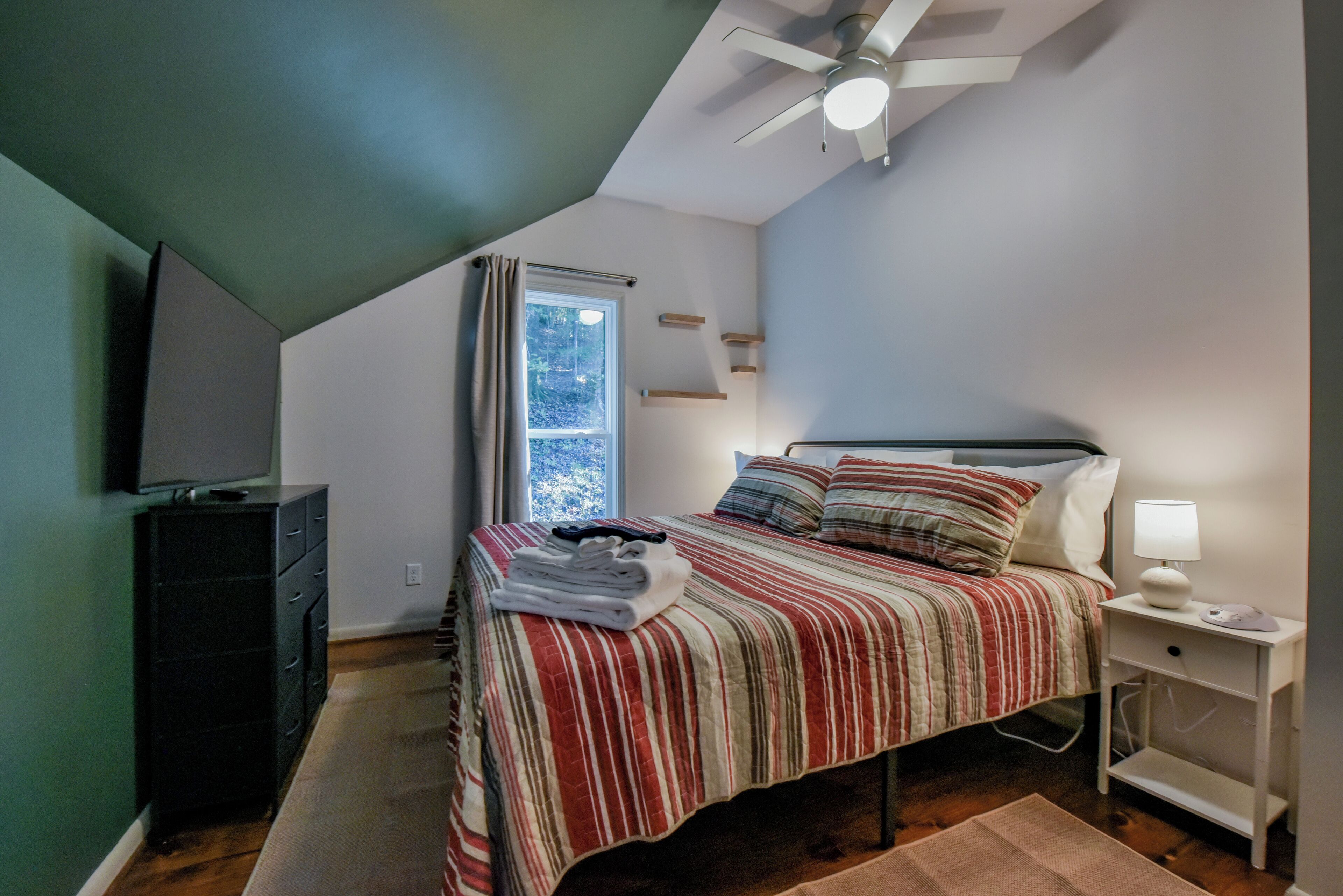 A bedroom with a bed, a nightstand, a TV, a window, and a ceiling fan.