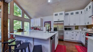 A modern kitchen with white cabinets, a large island with bar stools, stainless steel appliances, and large windows. - Legends Lodge: A woodland retreat near University of Alabama! (Cottondale)
