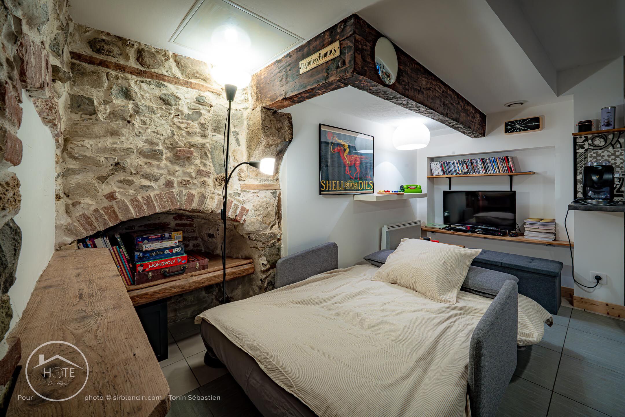 Une pièce chaleureuse avec un mur en pierre, un banc en bois avec des livres, un lit, une télévision et une étagère remplie de DVD.