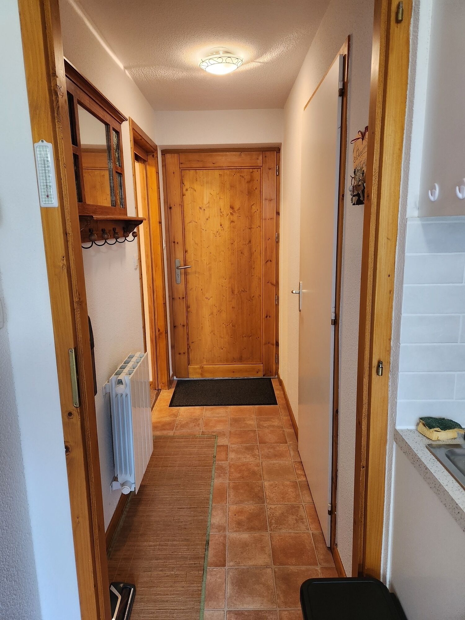 A narrow hallway with a wooden door at the end, a radiator on the left, and a tiled floor.