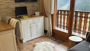 A cozy living area with a flat-screen TV, a wooden dining table, a sofa, and a view of a forested mountain through a balcony door. - 2 bedroom appt 20 mins from Morzine and rest of Portes du Soleil (Le Biot)