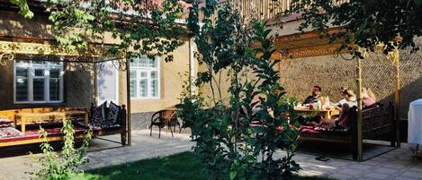Un espacio al aire libre con un banco y una mesa de madera, rodeado de vegetación y un edificio con paredes decoradas.
