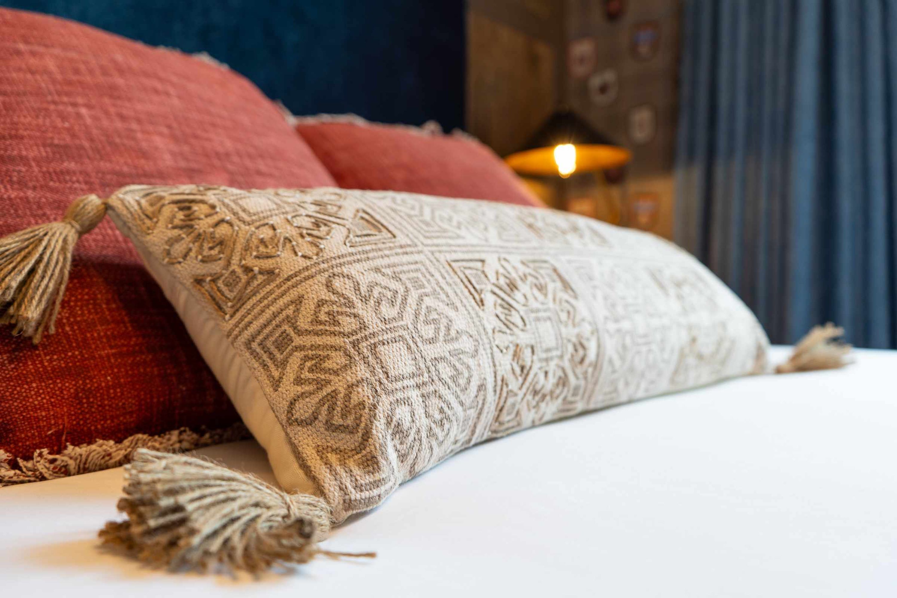 Una cama con un cojín estampado y flecos, un cojín rojo, y una vela encendida al fondo.
