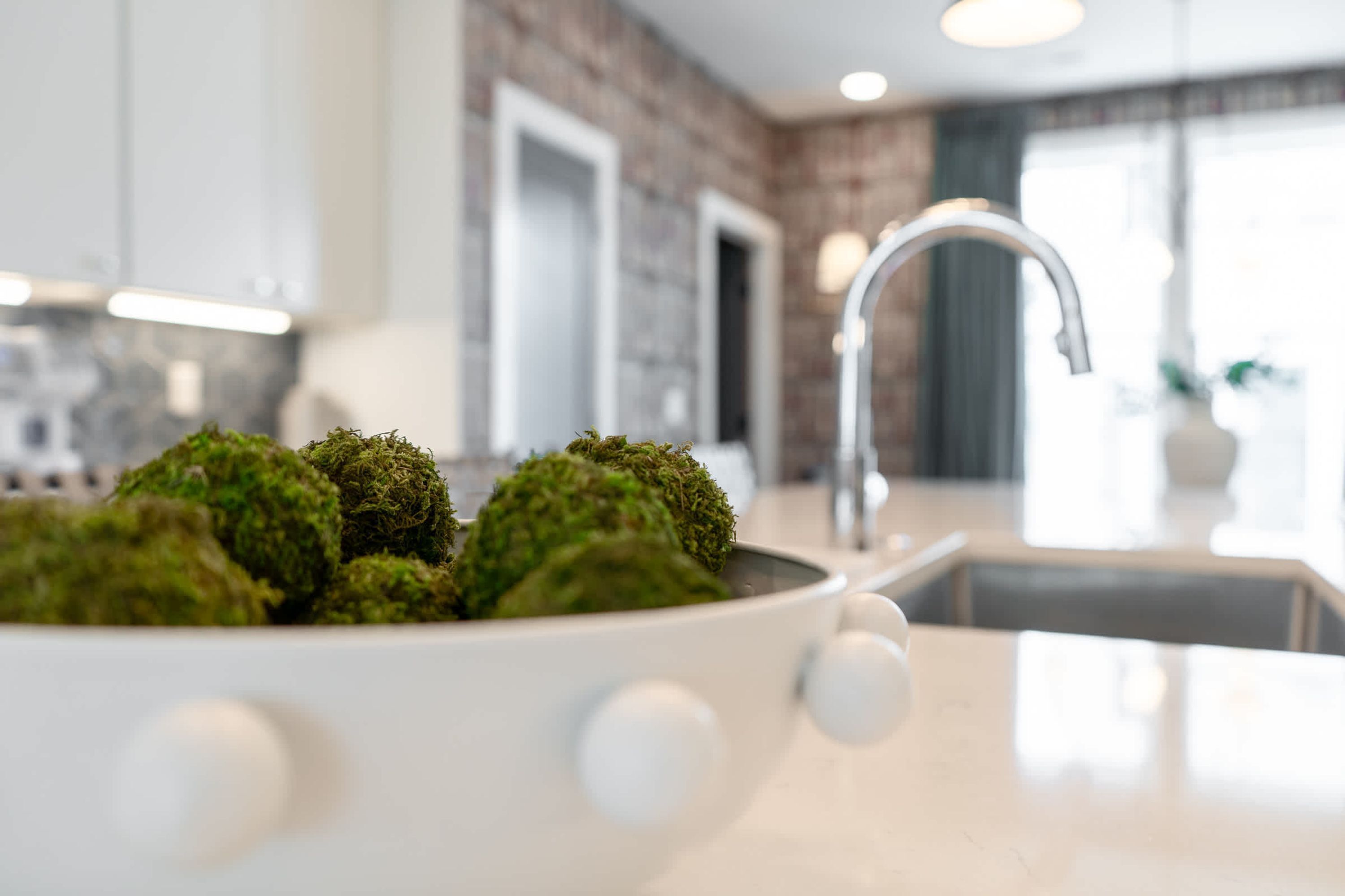 A modern kitchen with a white sink, a bowl of green moss, and a brick wall in the background.