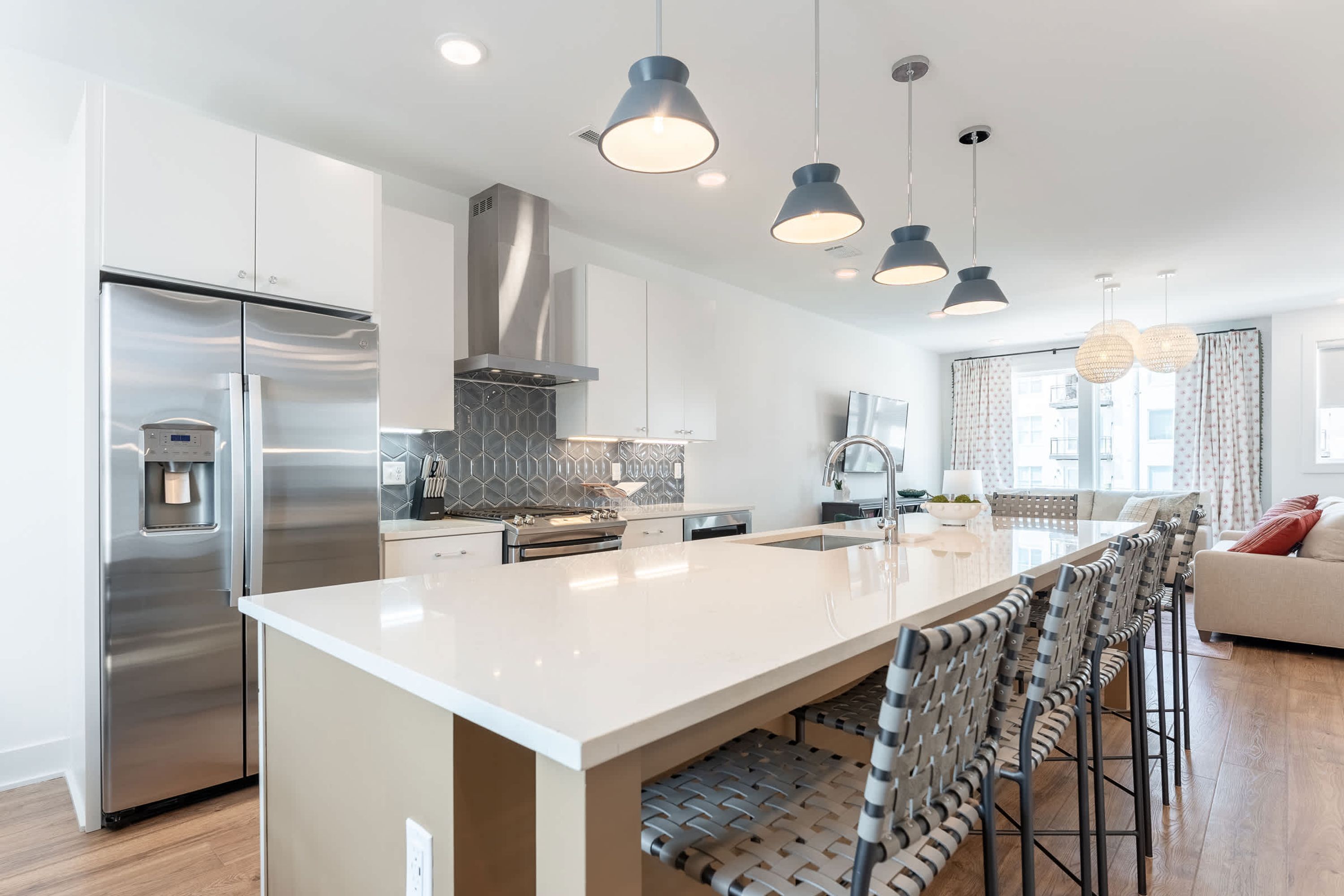 A modern kitchen with a stainless steel refrigerator, a large island with bar stools, pendant lighting, and a view to the living area.