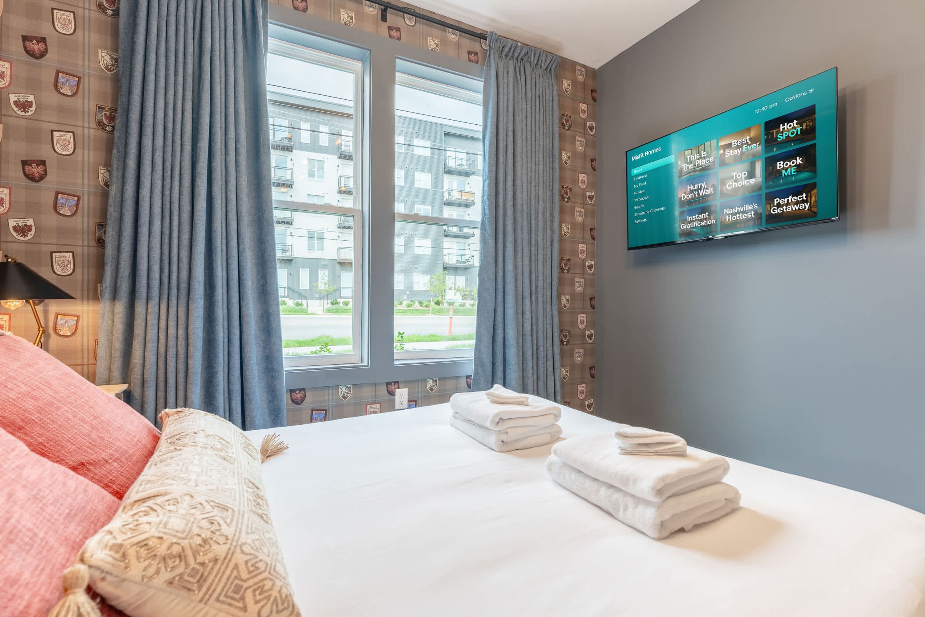 A hotel room with a bed, curtains, a TV mounted on the wall, and a window with a view of buildings.