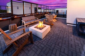Eine Dachterrasse mit Sitzgelegenheiten im Freien, einem Feuerkorb und Blick auf die Skyline der Stadt im Abendlicht.