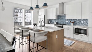 A modern kitchen with white cabinets, a central island, and a view of residential buildings. - Rooftop Fire Pit Lounge Skyline DT Nash Views (Nashville)