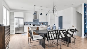 A modern kitchen with white cabinets, a stainless steel refrigerator, and a dining area with a dark table and striped chairs. - Rooftop Fire Pit Lounge Skyline DT Nash Views (Nashville)