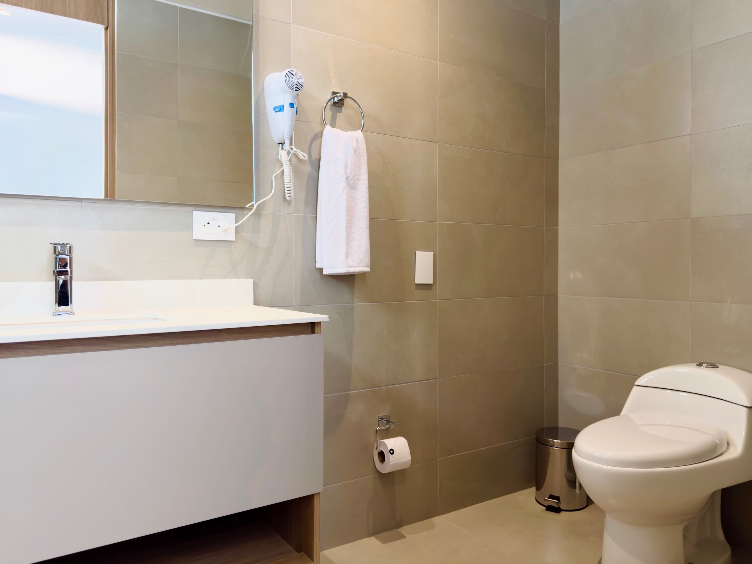 Une salle de bain moderne avec un lavabo blanc, un sèche-cheveux fixé au mur, des toilettes et une poubelle.