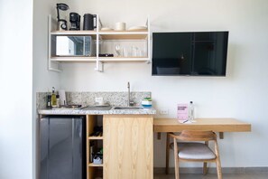 Une cuisine compacte équipée d’un réfrigérateur encastré, d’un four à micro-ondes et d’une machine à café posée sur une étagère. On y trouve une table en bois avec une chaise et un panneau posé dessus.