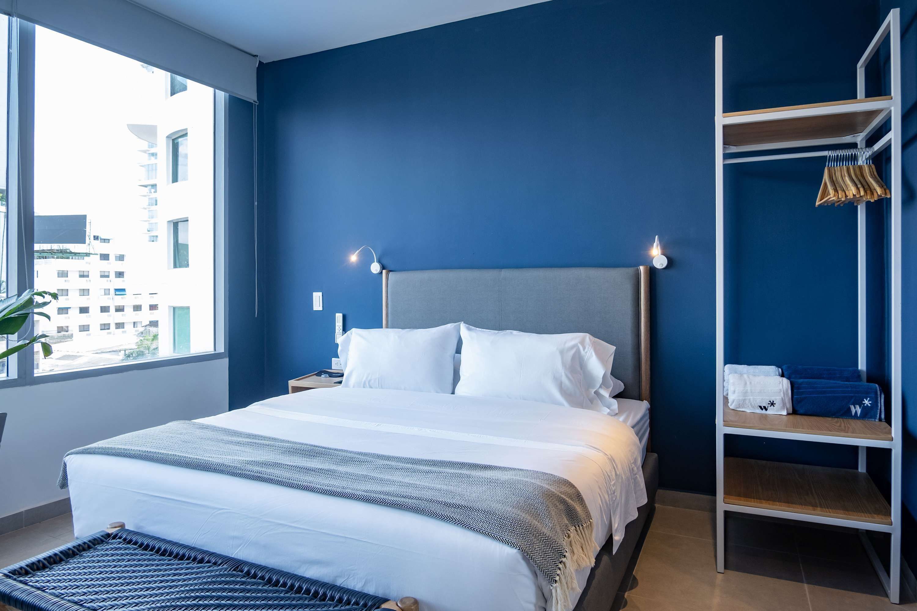 A modern hotel room with a large bed, a bedside table, a nightstand, a wardrobe, and a view of the city.