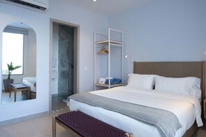 Une chambre d’hôtel moderne dotée d’un grand lit, d’un coin salon avec un banc, d’un dressing et d’une salle de bain avec une porte en verre.