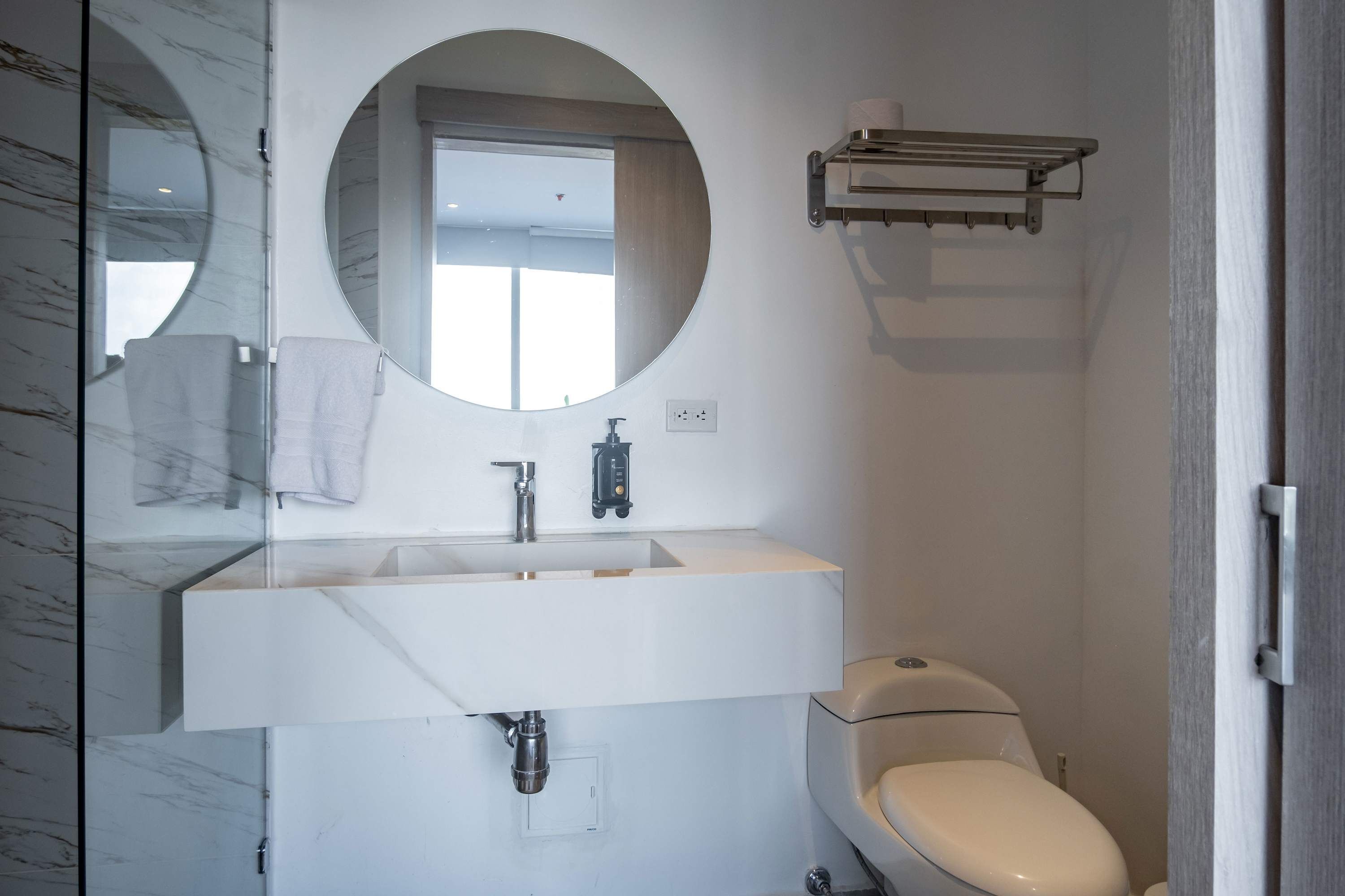 A modern bathroom with a marble vanity, a round mirror, a toilet, and a towel rack.