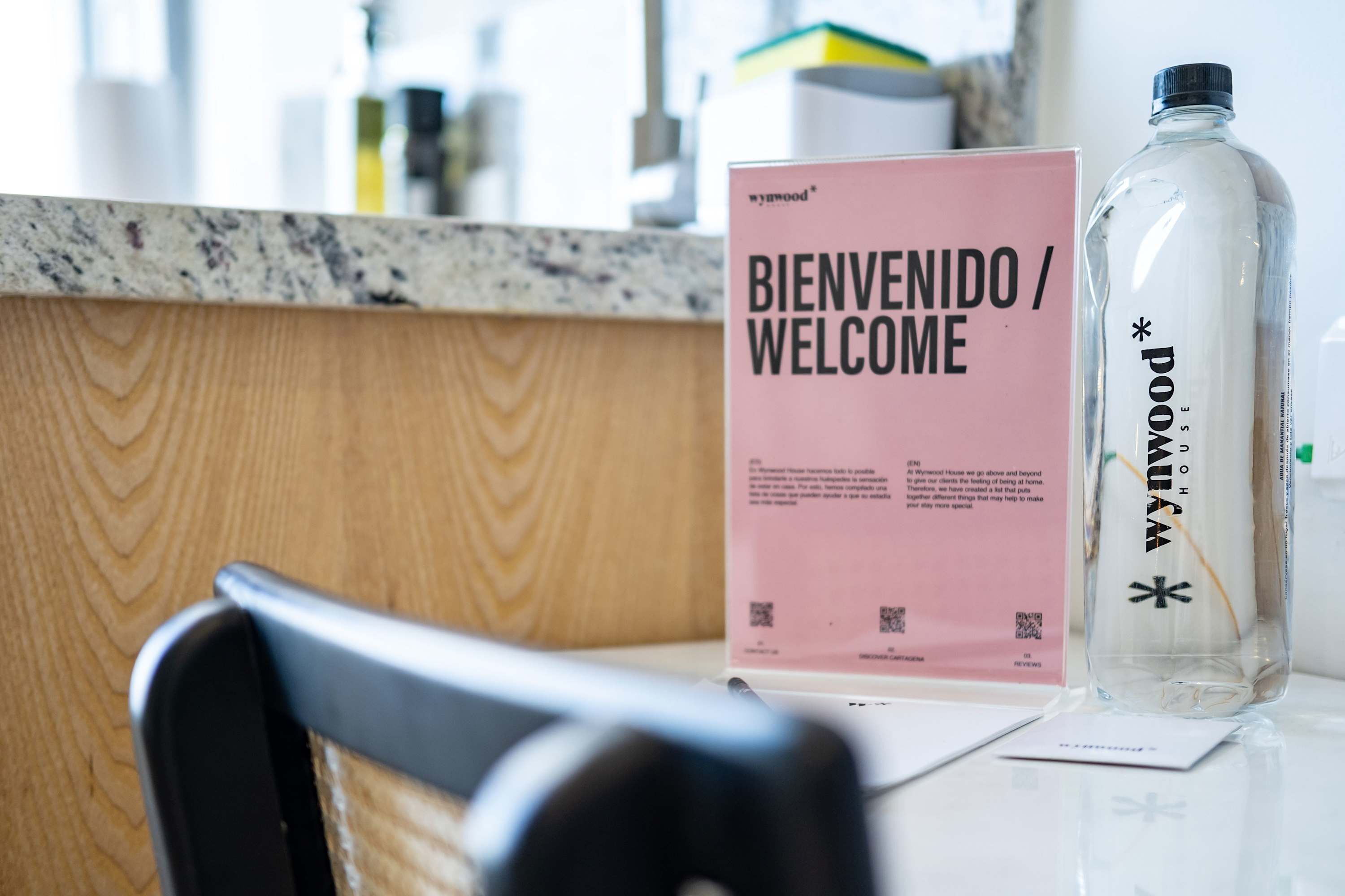Un comptoir d’accueil avec une pancarte indiquant « BIENVENIDO / WELCOME », une bouteille en verre transparent avec une étiquette et une chaise noire.