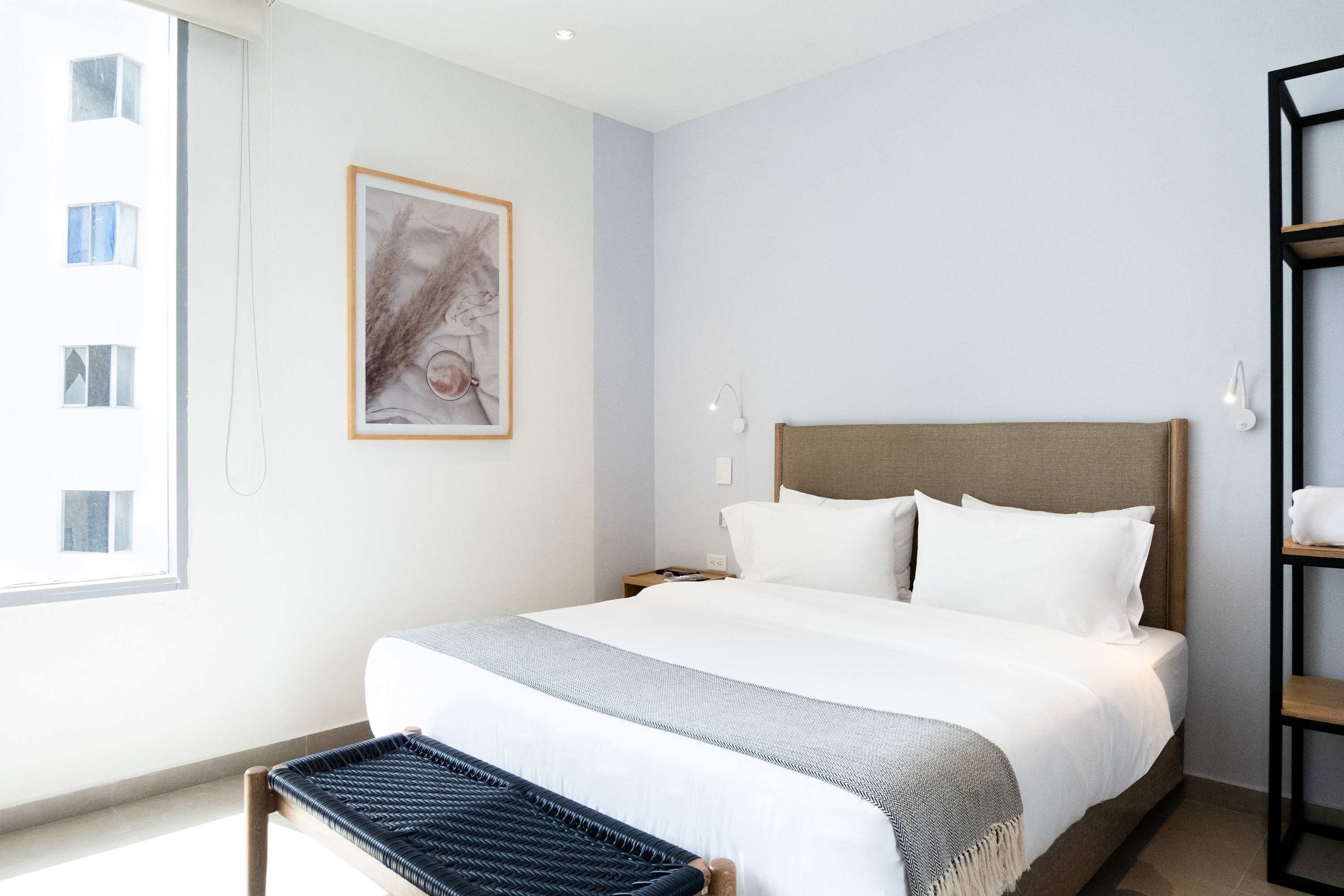 A hotel room with a large bed, a nightstand, a wall-mounted shelf, and a window with a view of buildings.