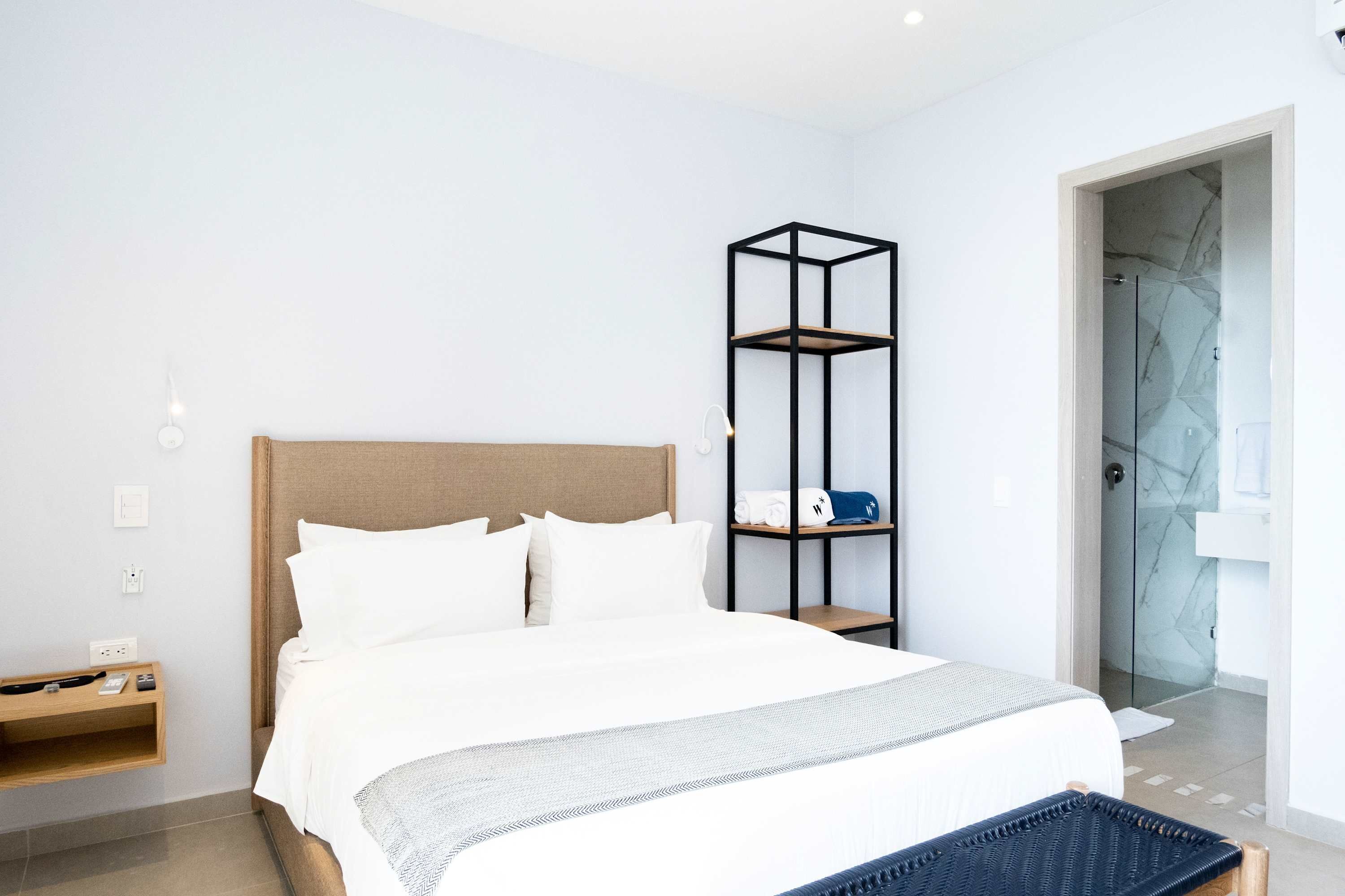 Una habitación de hotel moderna con una cama grande, una mesita de noche, un estante negro alto y una puerta de ducha de cristal.