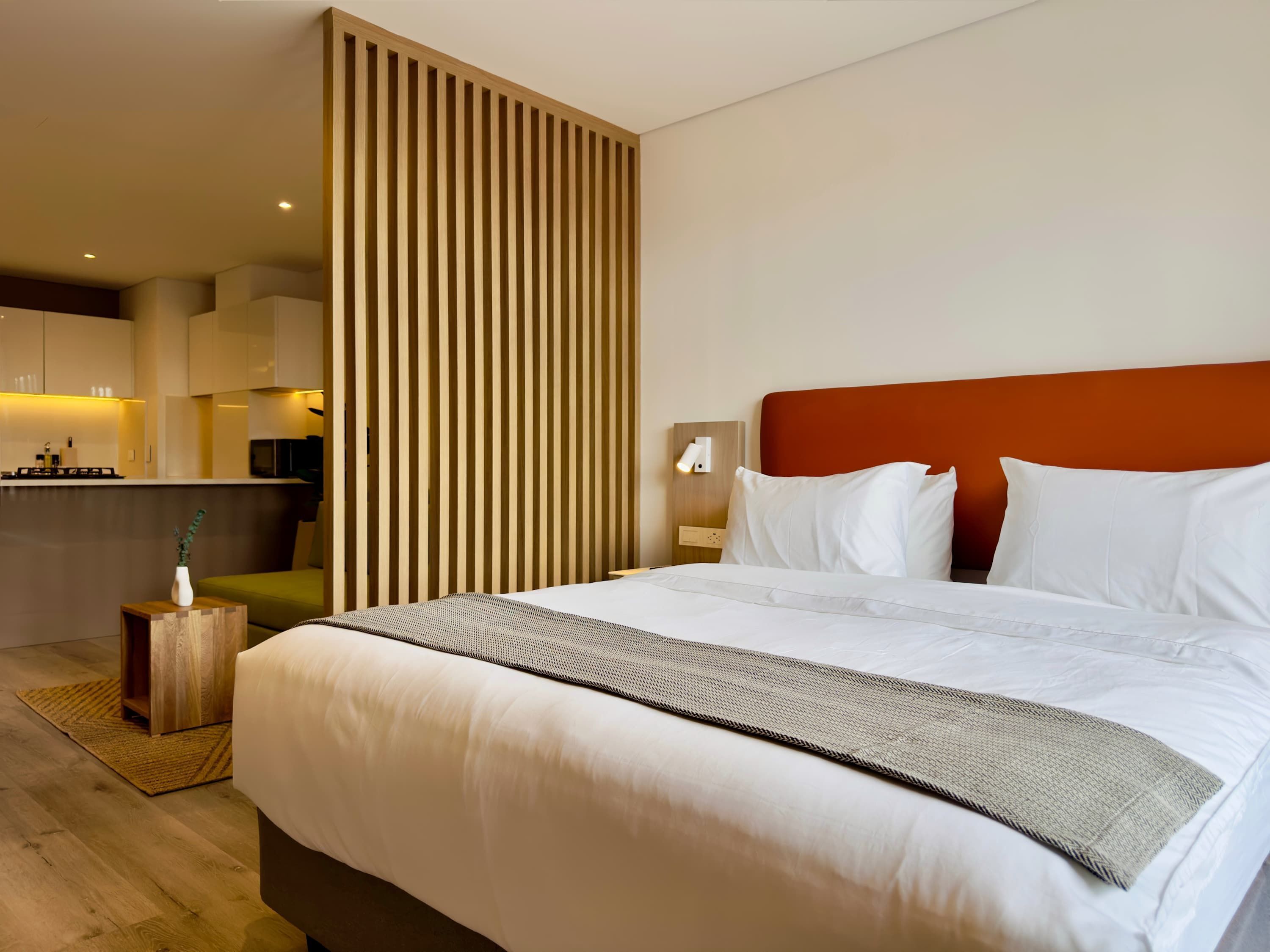 A modern hotel room with a large bed, a wooden partition, a kitchenette, and a view of the city.