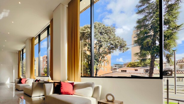 A modern hotel room with large windows, a sofa, and a small wooden side table. - w Cozy 2BR w Balcony in Santa Barbara (Bogotá)