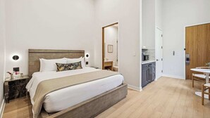 Quarto de hotel moderno com uma cama grande, uma mesinha de cabeceira, uma pequena escrivaninha e uma cozinha compacta.