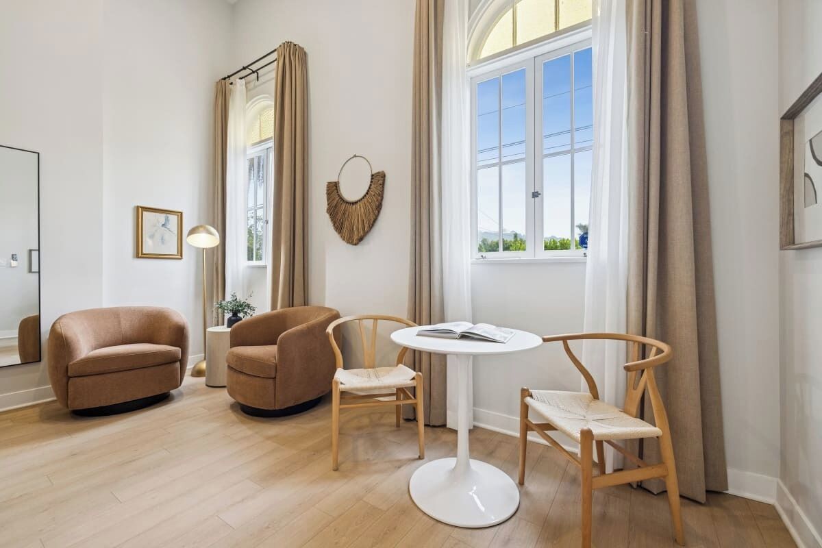 Un salon moderne avec une table ronde blanche, deux chaises en bois, un canapé marron et une grande fenêtre avec des rideaux.