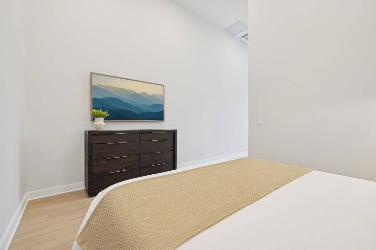 A bedroom with a bed, a dark dresser, a painting of mountains, and a small plant on the dresser.