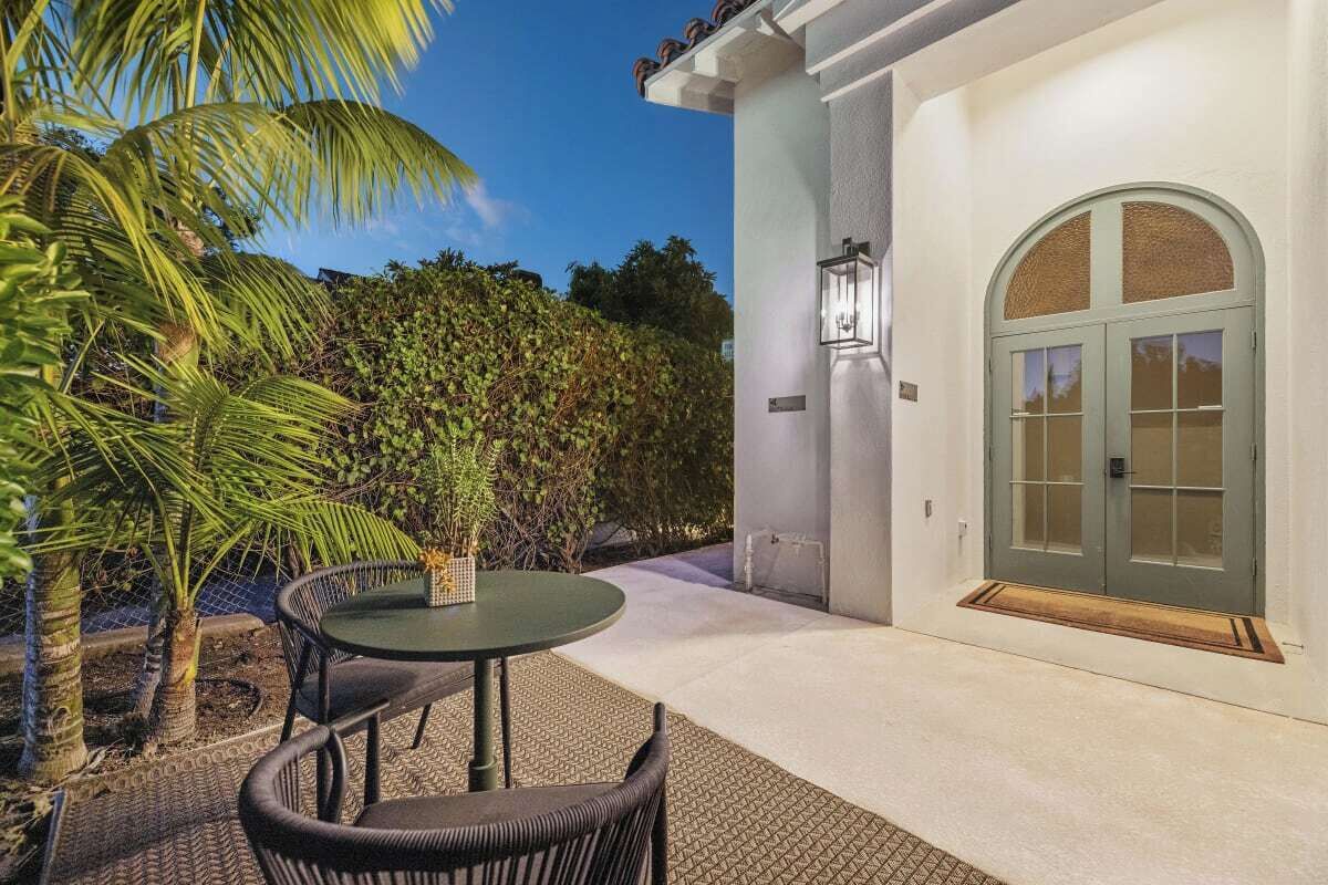 An outdoor patio with a round table and chairs, a palm tree, and a white building with a green door.